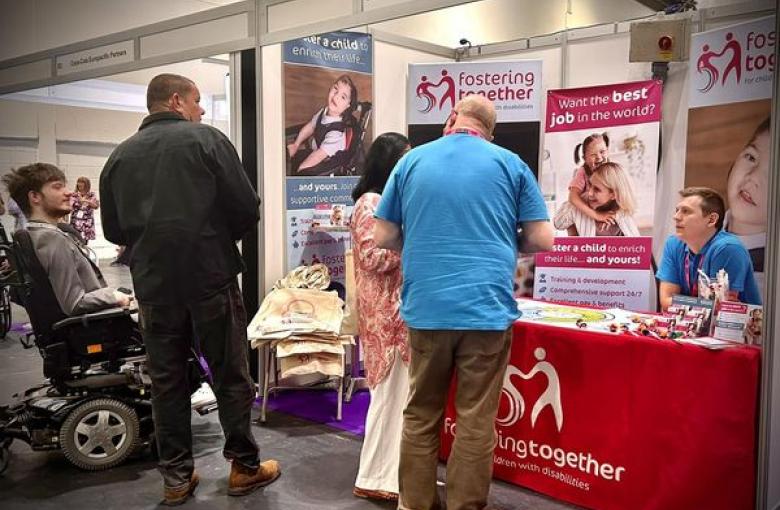 The Fostering Together team at the Disability Expo 2023. The image shows two people looking at our material whilst Neil and Jack from Fostering Together are also chatting to another person at the stand. The stand has lots of information on banners and on the table and the table is coverred with a cloth with Fostering Together's signature pink and the heart logo.